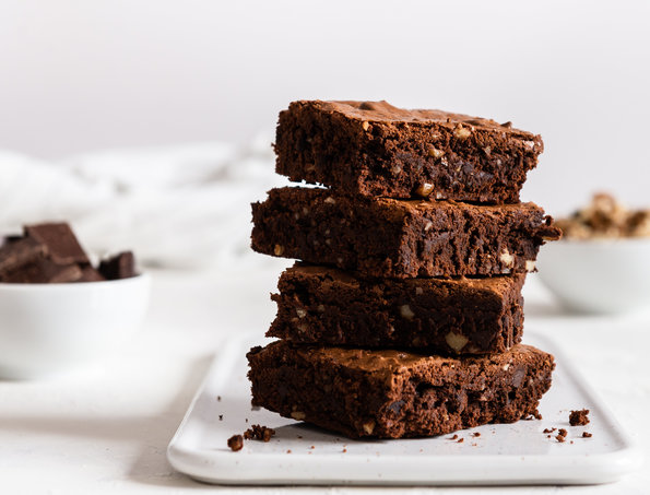 Stack of four chocolate brownies on a white plate, with behind them bowls of chocolate and nuts on a white background